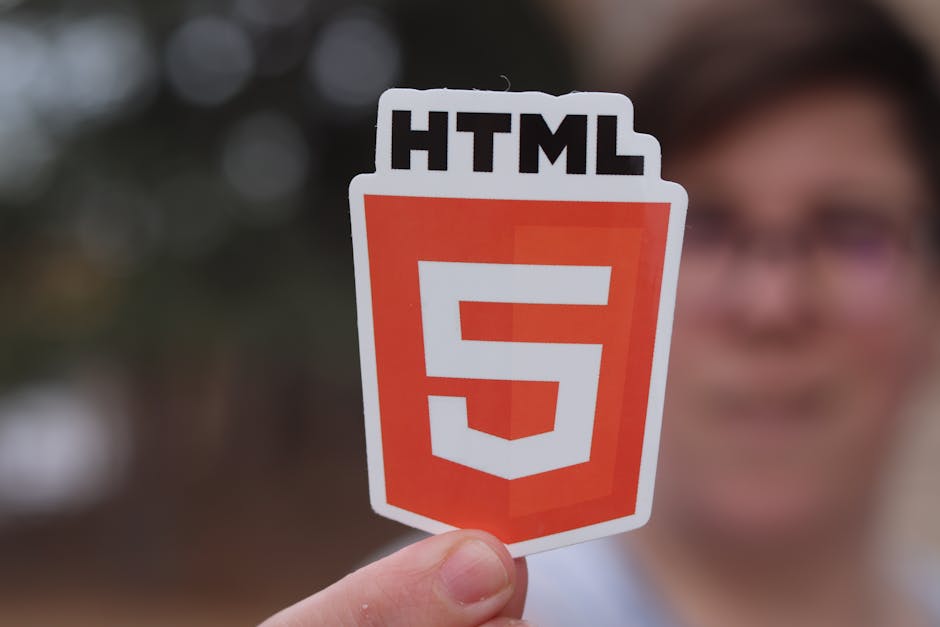 A person holds a small cutout sticker featuring the HTML5 logo, which consists of a shield shape with a red-orange background and a large white number '5' in the center, along with the text 'HTML' at 