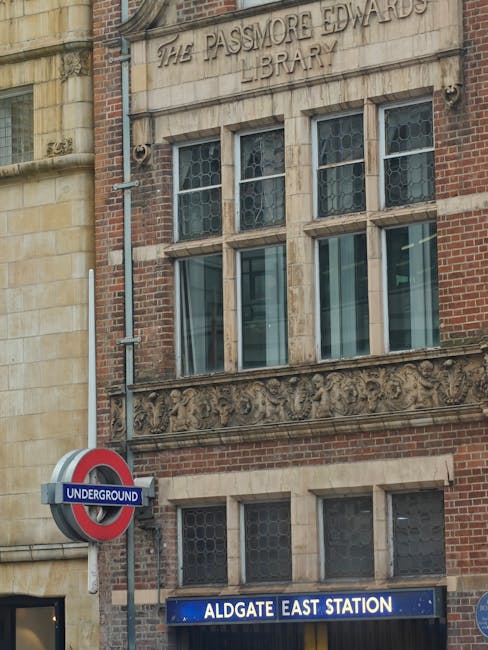 A narrow urban street scene showing a mix of historic and modern buildings, including a prominent red-brick structure with large, arched windows and ornate architectural details, topped with a teal-co